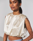 A woman wearing a sleeveless champagne-colored satin top with a textured dotted pattern and a cropped, gathered silhouette, paired with light-wash blue jeans against a plain light gray background.