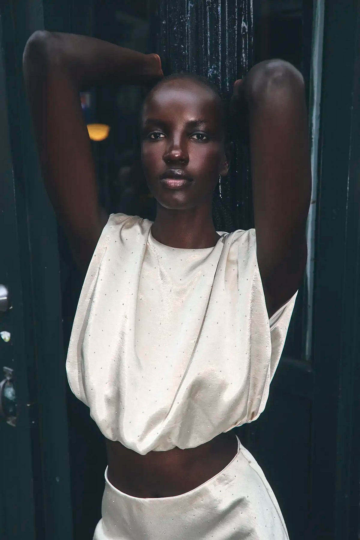 A model with a shaved head poses against a dark green door, wearing a cream-colored, textured two-piece set consisting of a cropped top and matching skirt.