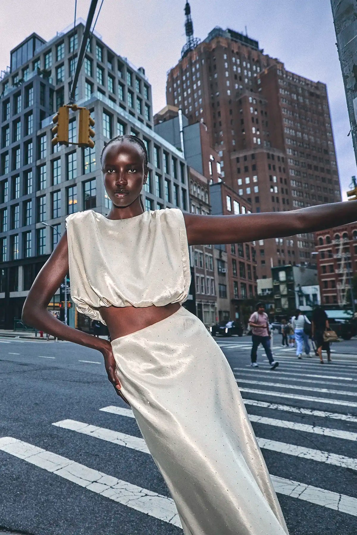 A model wearing a shimmering cream satin two-piece set consisting of a cropped puff-sleeve top and a matching high-waisted skirt, posing on a city crosswalk with skyscrapers in the background.