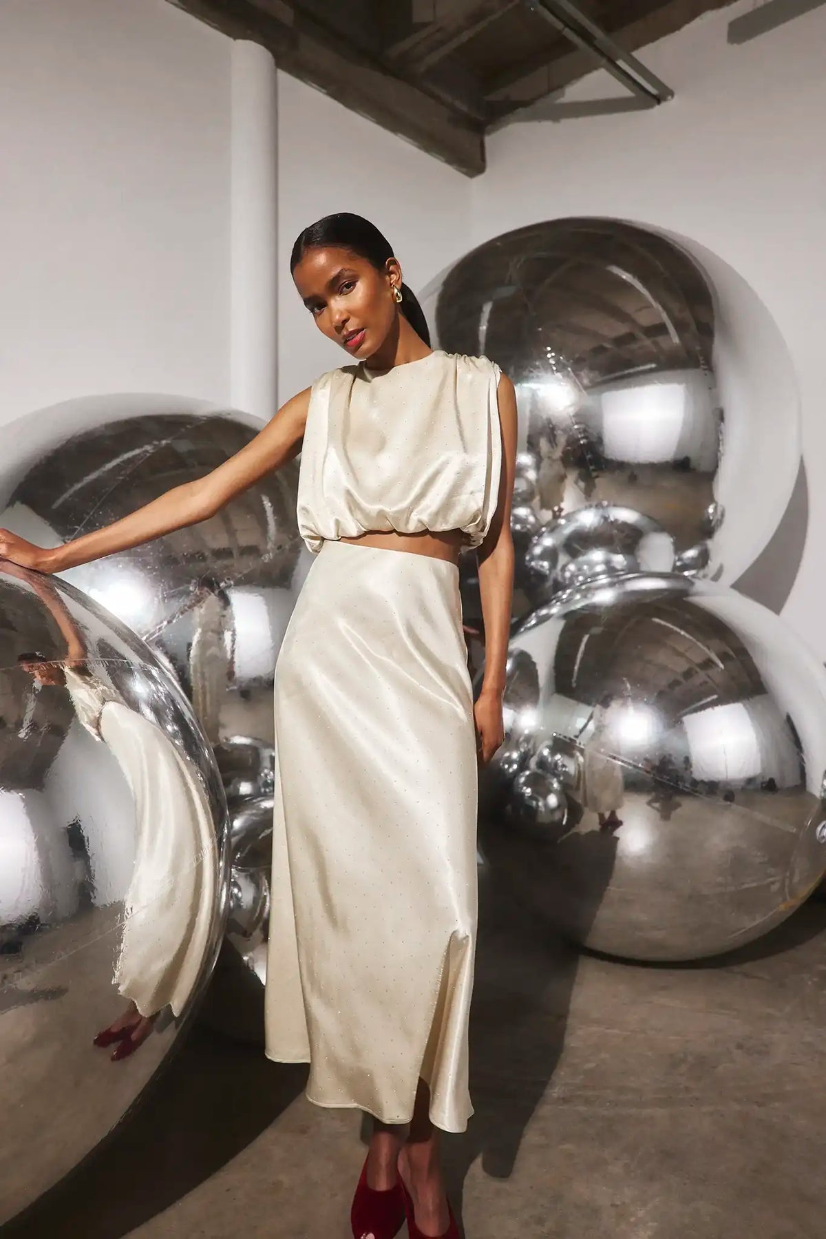 A model wearing a cream satin two-piece set consisting of a cropped sleeveless top and a matching high-waisted skirt, posing in front of large reflective silver mirrors in an art gallery setting.