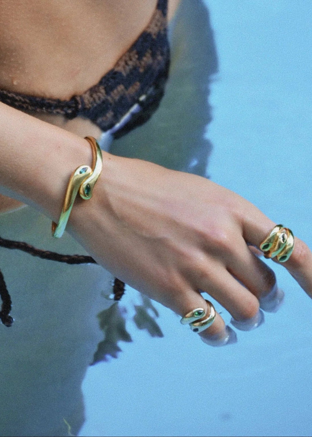 A close-up lifestyle shot of a person's hand and wrist wearing gold snake-themed jewelry, including a wrap-around bracelet and matching rings, set against a blue water background.