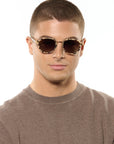 A man with a buzz cut wearing round tortoiseshell sunglasses and a brown knit sweater, photographed against a plain white background.