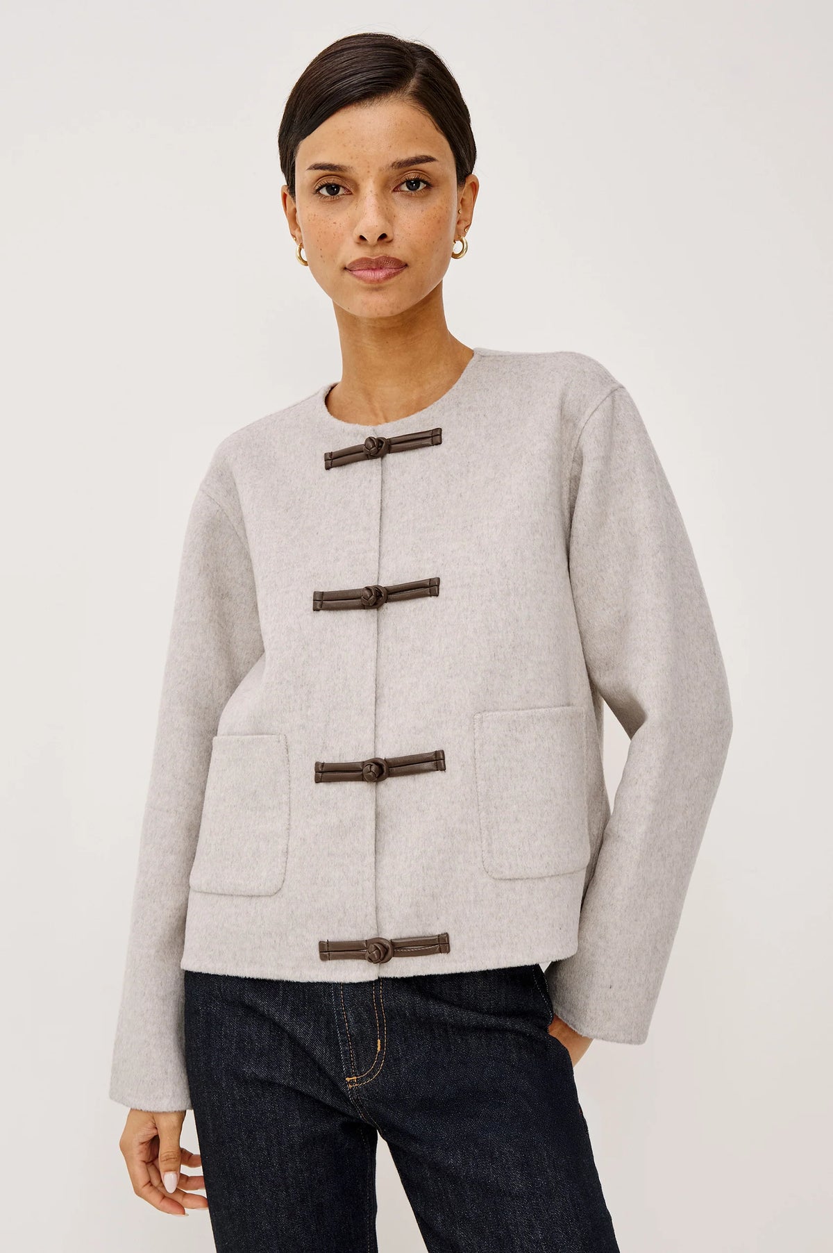 model wearing an oat colored jacket with toggle closures and jeans on a light grey background