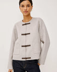 model wearing an oat colored jacket with toggle closures and jeans on a light grey background