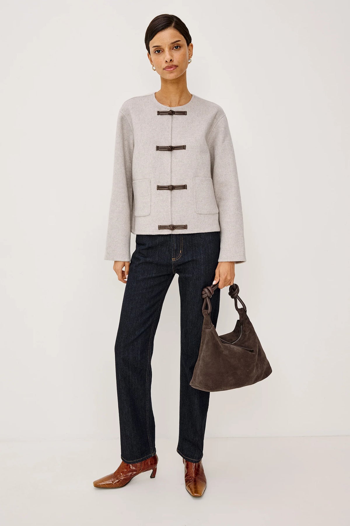 model wearing an oat colored jacket with toggle closures and jeans on a light grey background