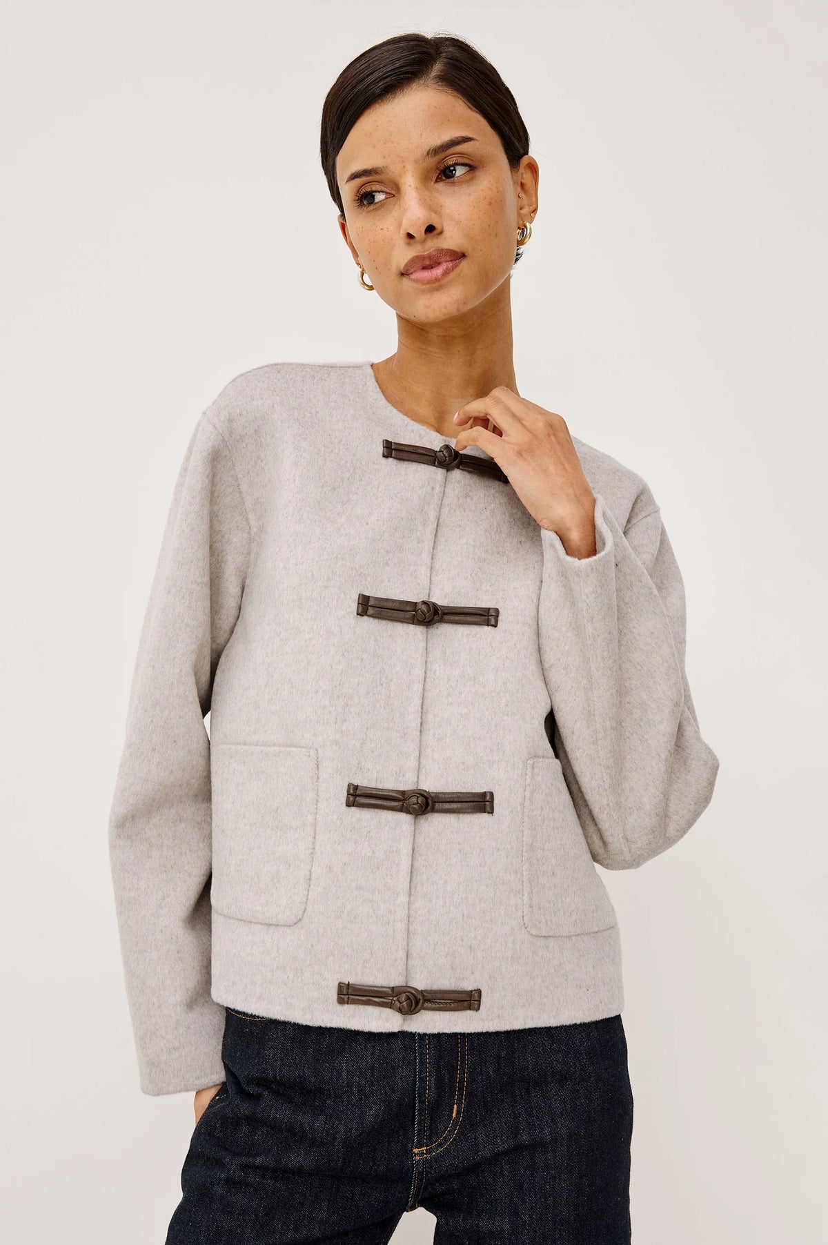 model wearing an oat colored jacket with toggle closures and jeans on a light grey background