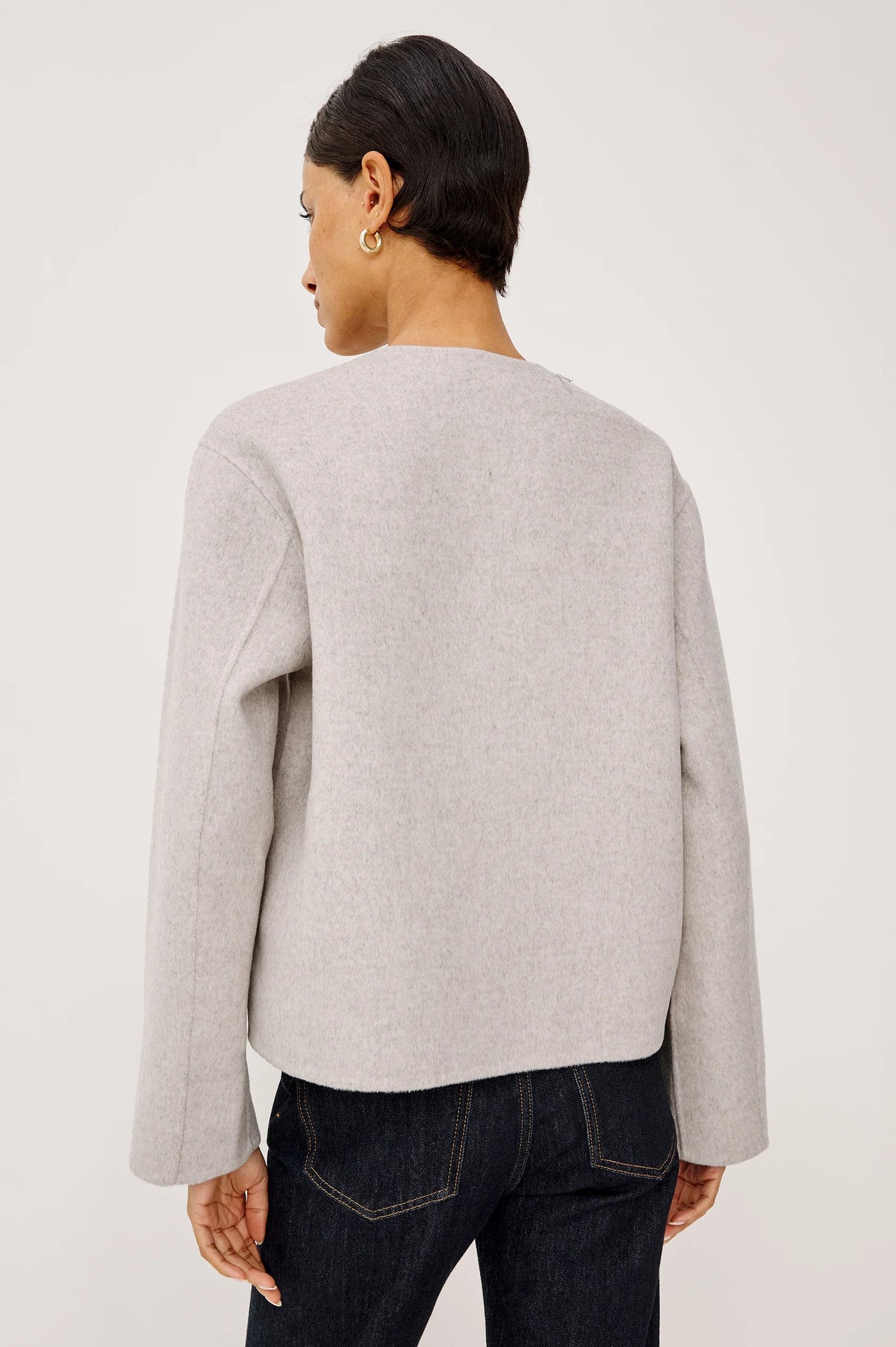 model wearing an oat colored jacket with toggle closures and jeans on a light grey background, back view
