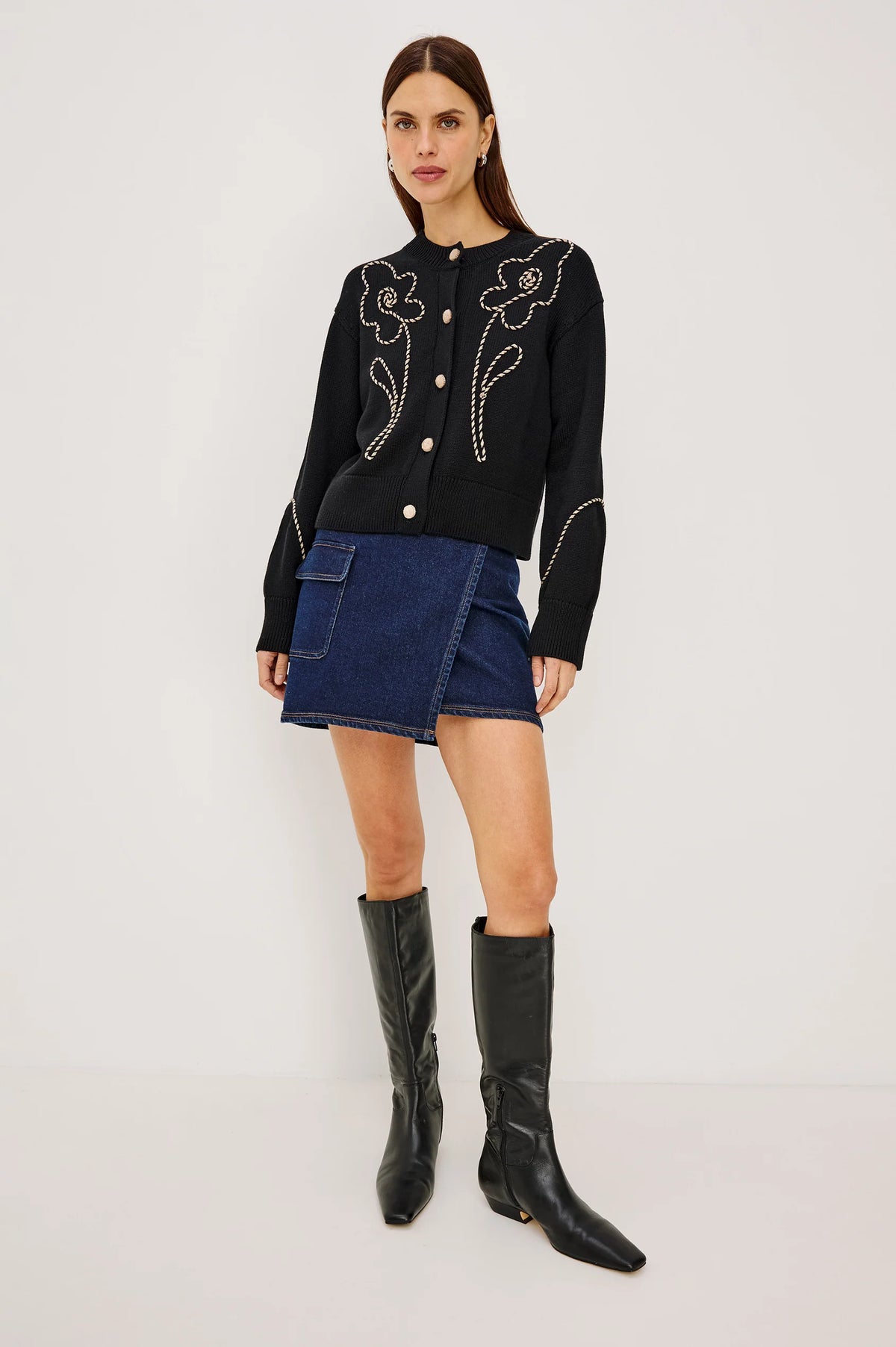 A model stands against a plain light gray studio background wearing a black long-sleeve knit top with white floral embroidery and gold-tone buttons, a dark blue denim mini skirt, and black knee-high leather boots.