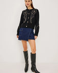 A model stands against a plain light gray studio background wearing a black long-sleeve knit top with white floral embroidery and gold-tone buttons, a dark blue denim mini skirt, and black knee-high leather boots.