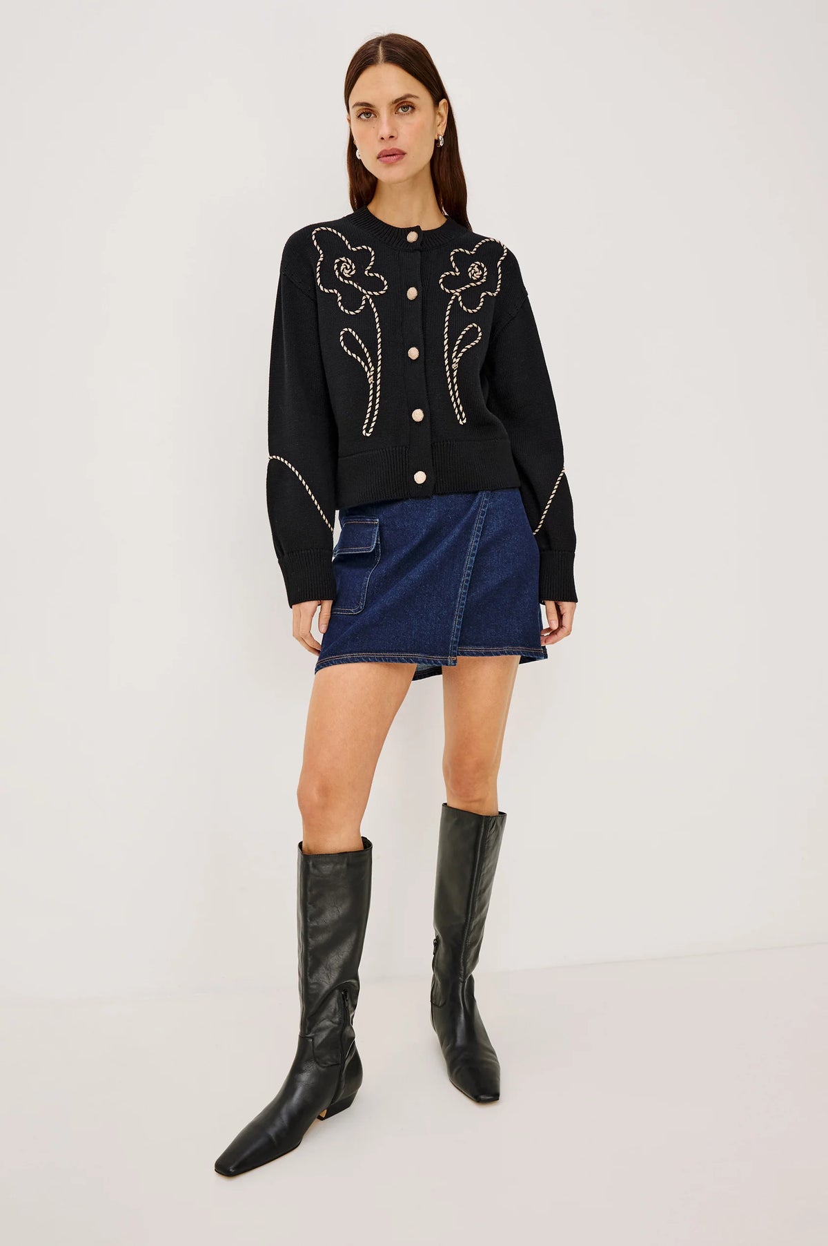 A model stands against a light gray studio background wearing a black long-sleeve knit top with white floral embroidery and gold buttons, a blue denim mini skirt, and black knee-high leather boots.