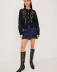 A model stands against a light gray studio background wearing a black long-sleeve knit top with white floral embroidery and gold buttons, a blue denim mini skirt, and black knee-high leather boots.