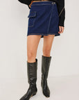 A model wearing a dark blue denim mini skirt with cargo pockets, styled with a black cropped jacket featuring silver chain details and black knee-high leather boots, set against a plain white studio background.