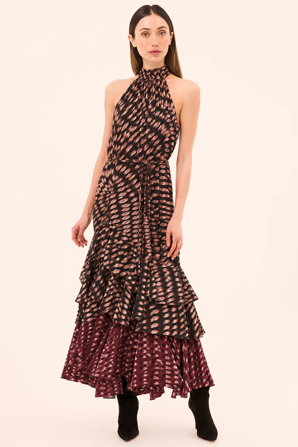 A model wearing a sleeveless halter-neck maxi dress with a black and beige abstract pattern on the bodice and a dark burgundy and beige patterned layered ruffle hem, paired with black ankle boots, against a solid light beige background.