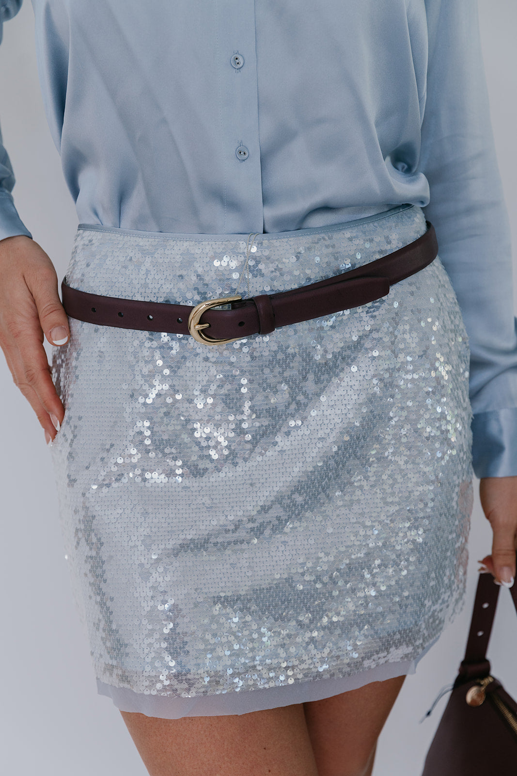 Person wearing a sequin skirt with a brown belt and holding a handbag on a plain background