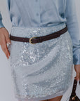 Person wearing a sequin skirt with a brown belt and holding a handbag on a plain background