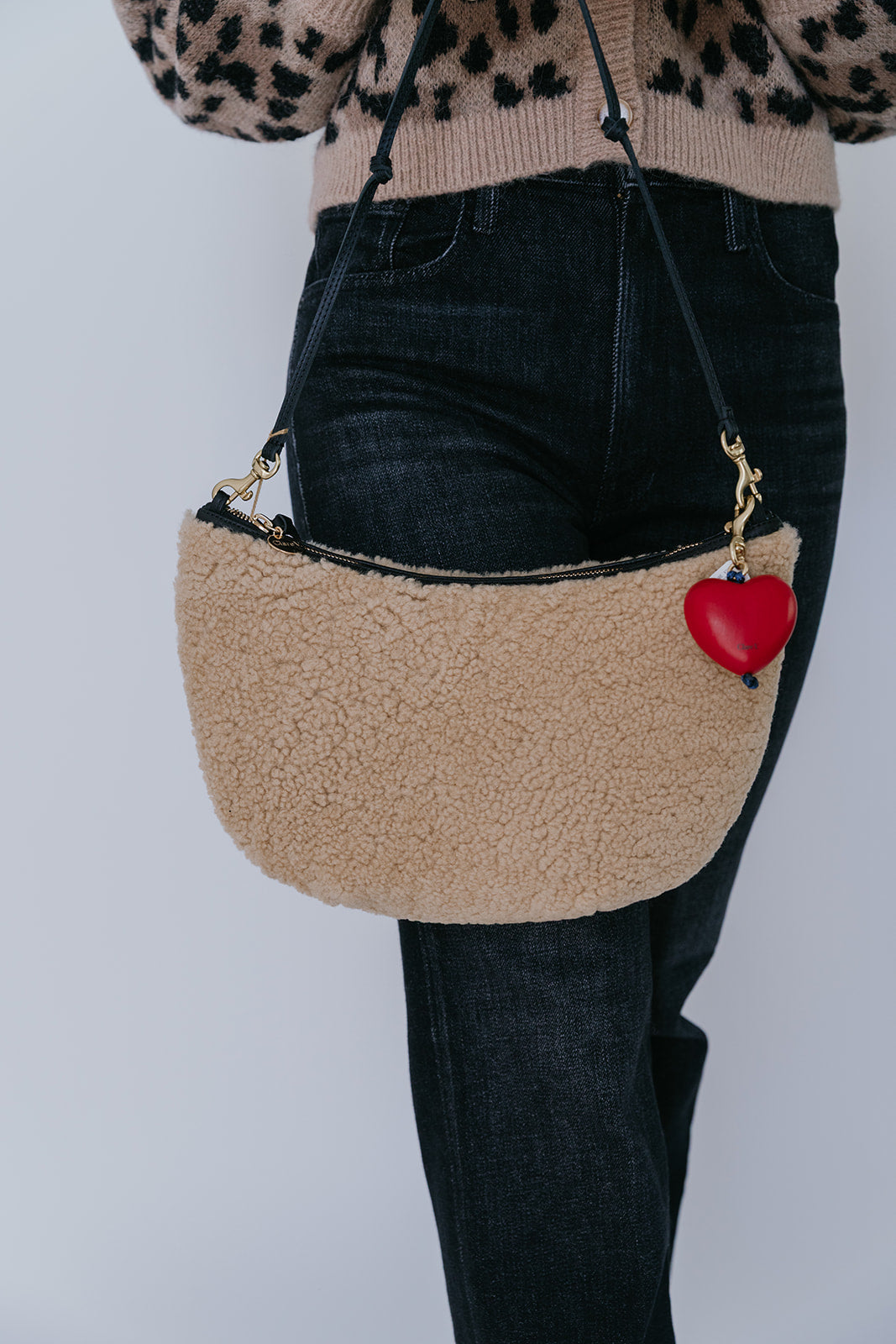 Beige handbag with a red heart charm worn by a person against a plain background