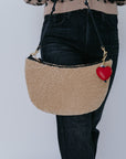 Beige handbag with a red heart charm worn by a person against a plain background
