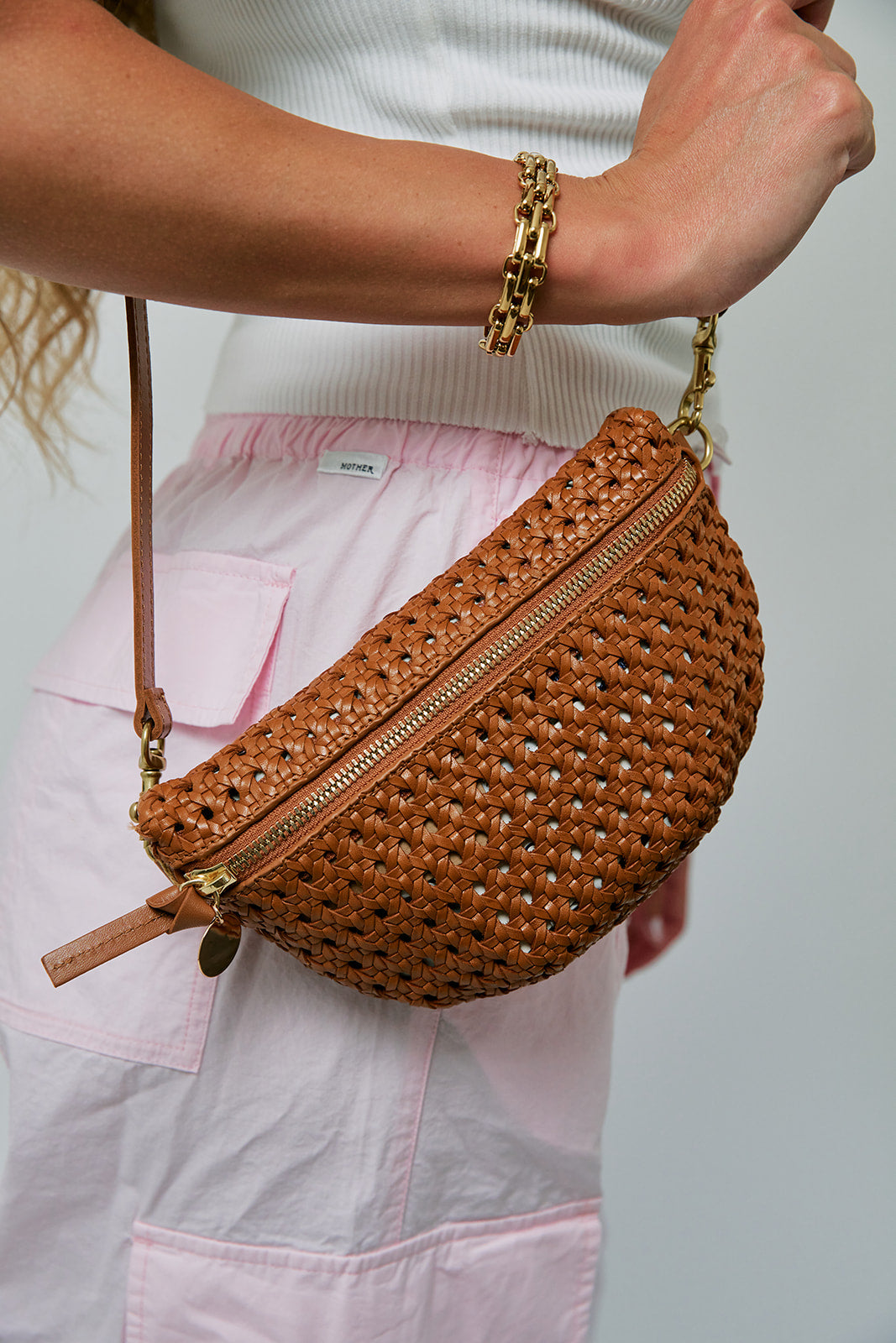 Brown woven handbag held by a person wearing a white top and pink pants.