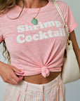 Person wearing a pink t-shirt with 'Shrimp Cocktail' text, holding a beige handbag.