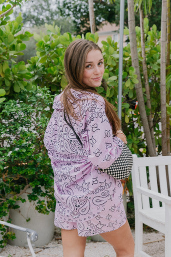Woman wearing a purple patterned dress standing outdoors with greenery in the background, back view