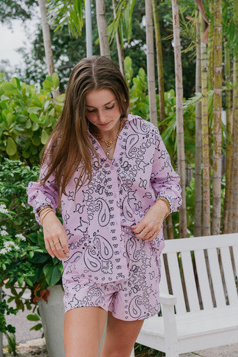 Woman wearing a purple paisley outfit standing outdoors with greenery in the background front view