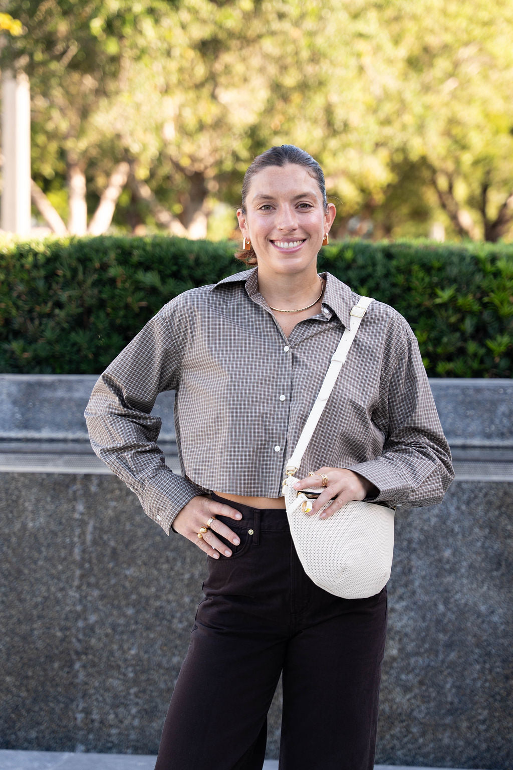 Woman standing outdoors with a white bag, wearing a checkered shirt and dark pants.