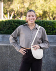 Woman standing outdoors with a white bag, wearing a checkered shirt and dark pants.