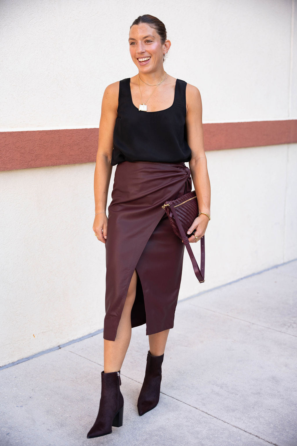Woman wearing a black top and brown skirt with a clutch against a white wall.