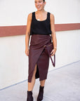 Woman wearing a black top and brown skirt with a clutch against a white wall.