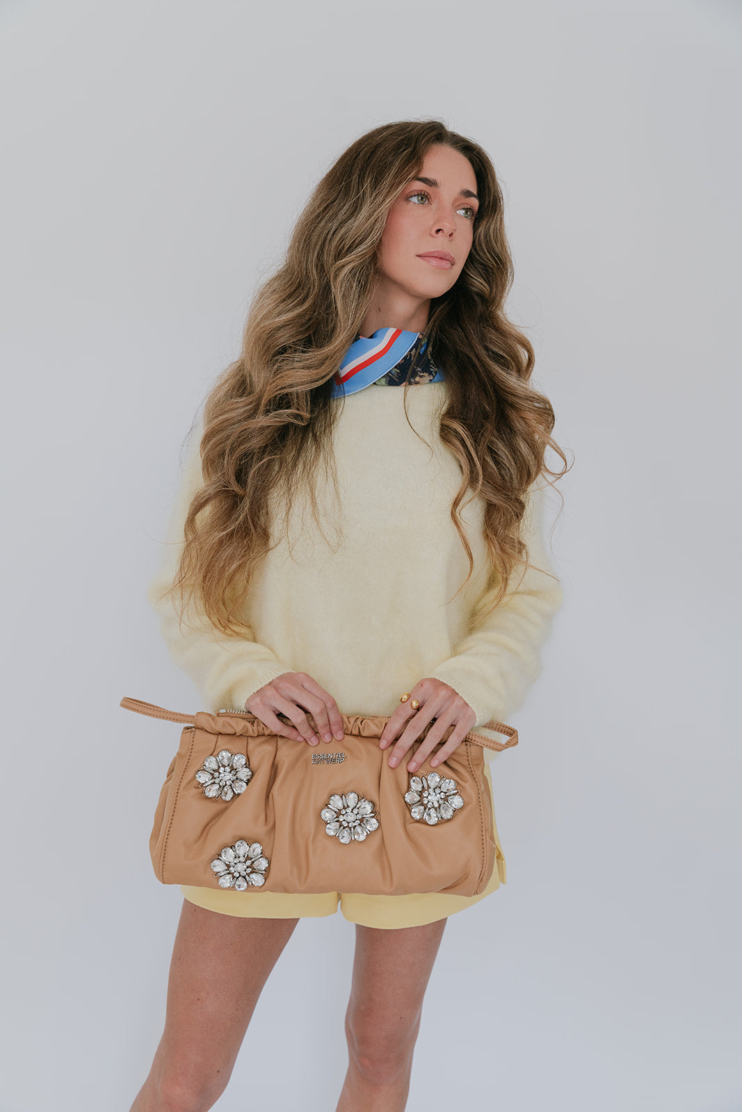 Woman wearing yellow sweater, holding a beige clutch with floral embellishments against a plain background