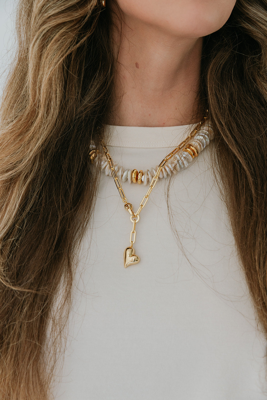 Close-up of a person wearing a gold necklace with a heart pendant and another necklace with beads.