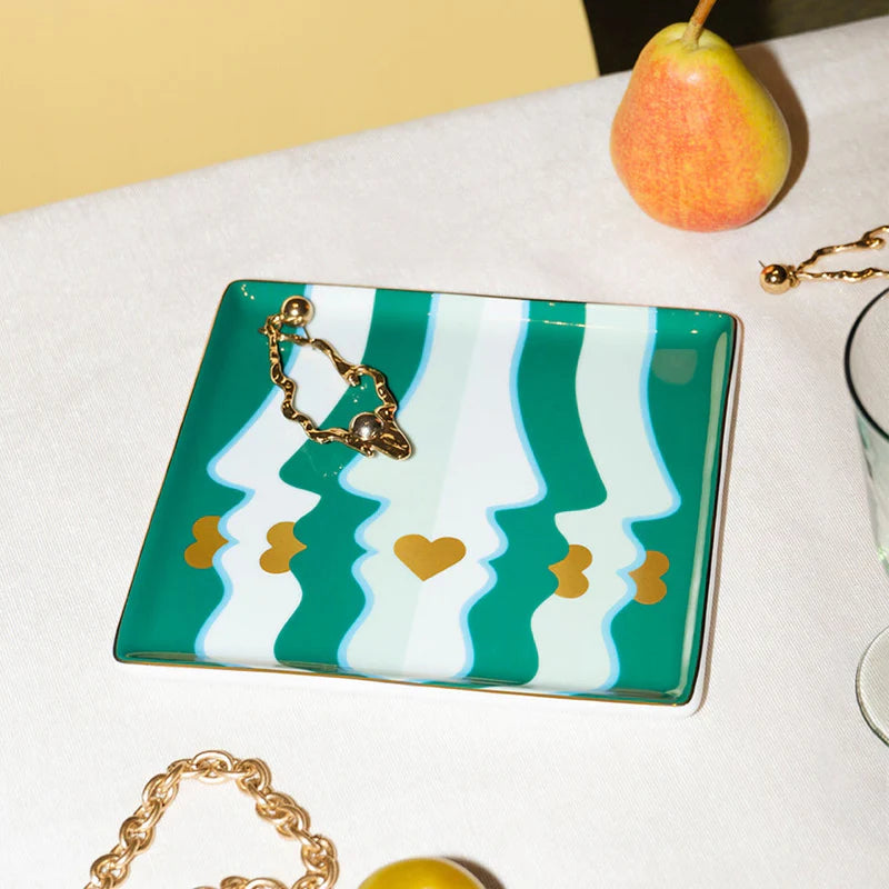 Rectangular clutch with green and white wave pattern and gold chain on a white surface with a pear and gold bracelet.