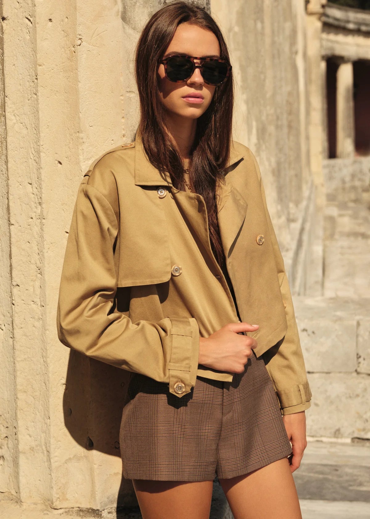Woman wearing a beige jacket and sunglasses standing against a stone wall.