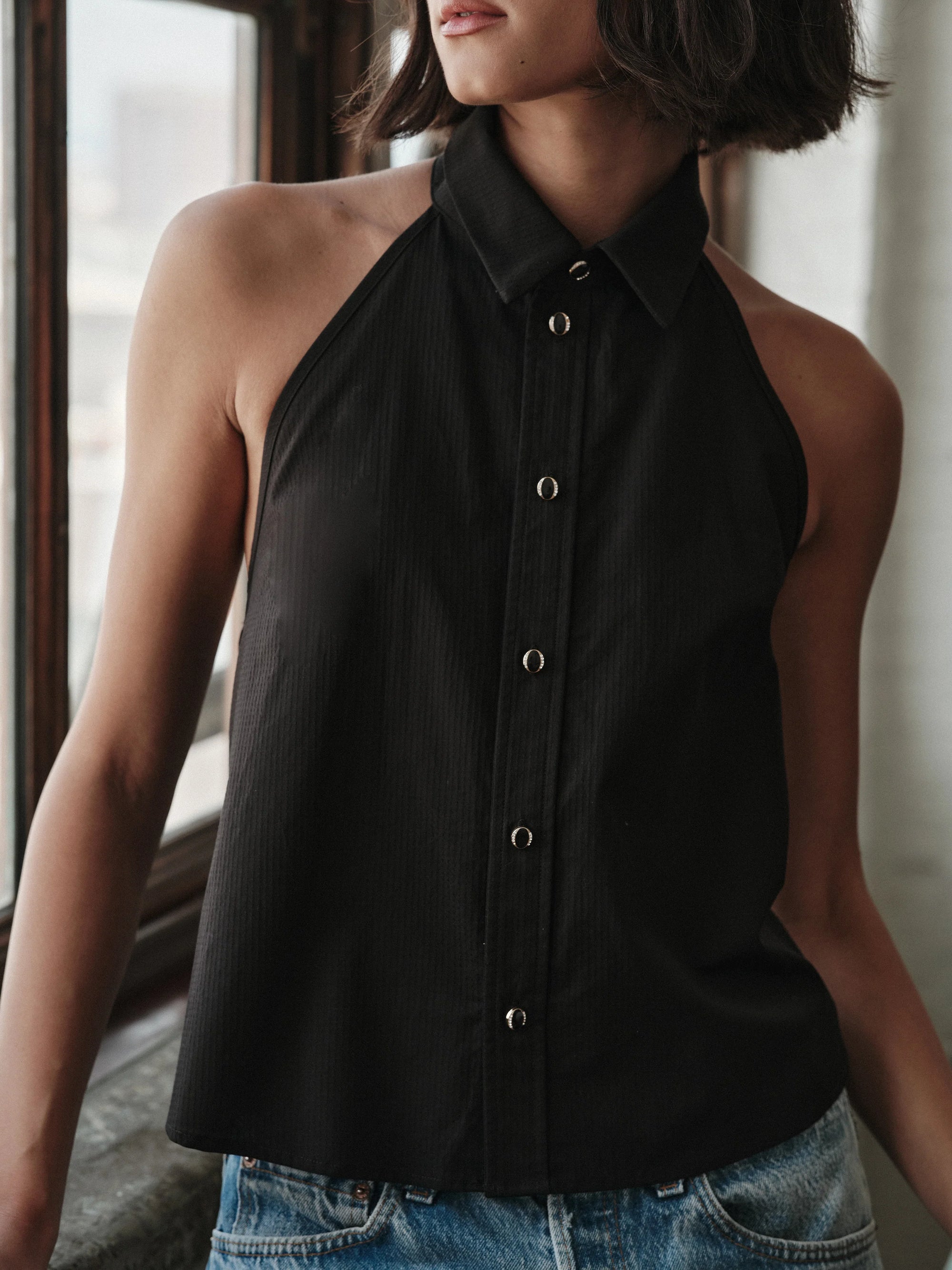 Woman wearing a black halter neck top with buttons and blue jeans indoors.