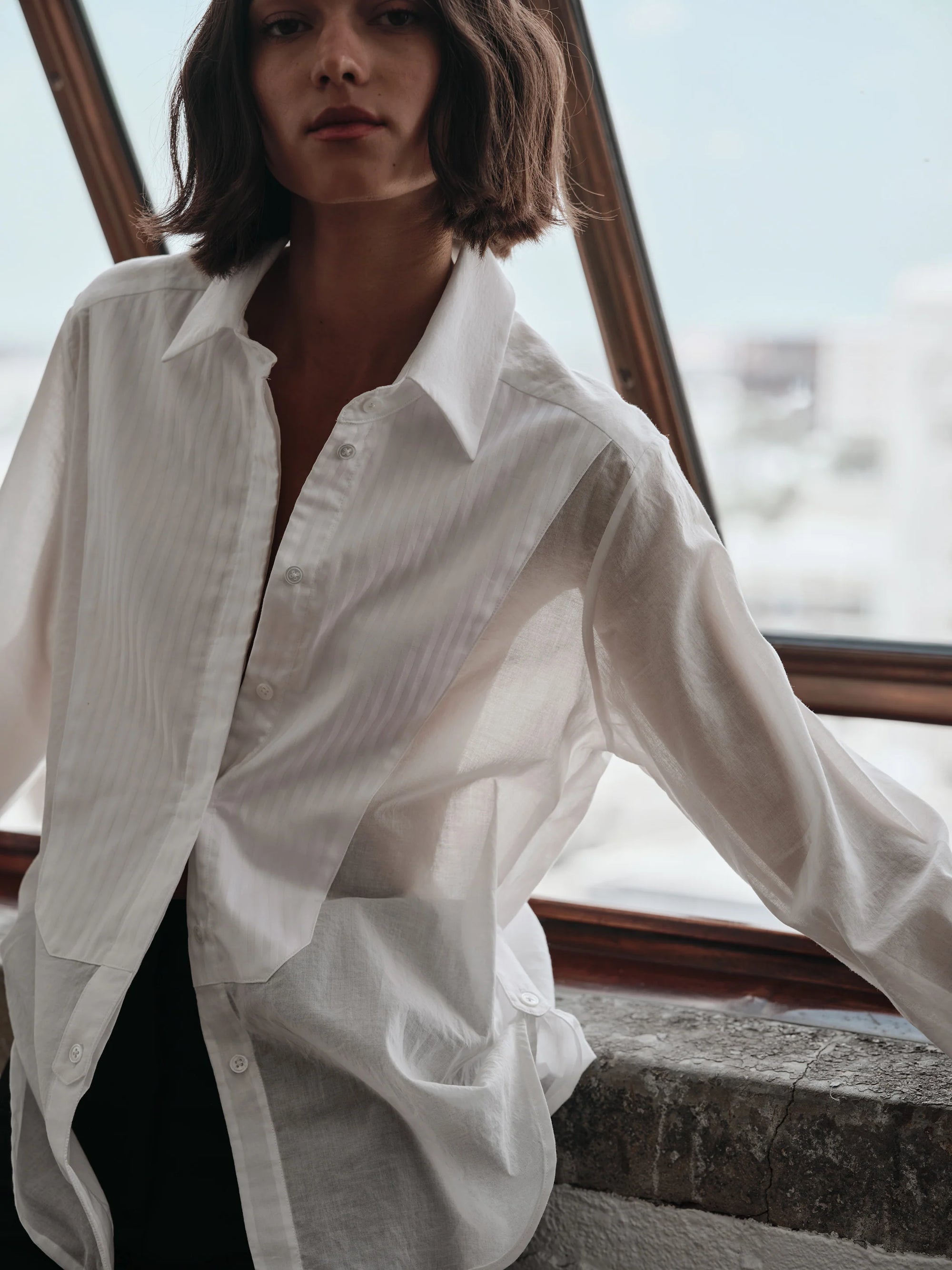 Person wearing a white shirt standing by a window with a blurred cityscape background