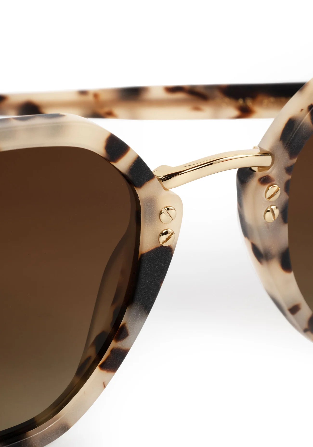 Close-up of a pair of sunglasses with a tortoiseshell patterned acetate frame, gold metal hinge, and dark brown gradient lenses on a white background.