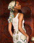Woman wearing a white sleeveless top and patterned skirt with a headscarf, standing against a wooden wall.