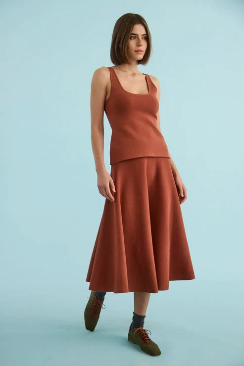A woman modeling a rust-orange two-piece knit set consisting of a sleeveless scoop-neck top and a matching A-line midi skirt, paired with dark socks and brown lace-up shoes, against a solid light blue studio background.