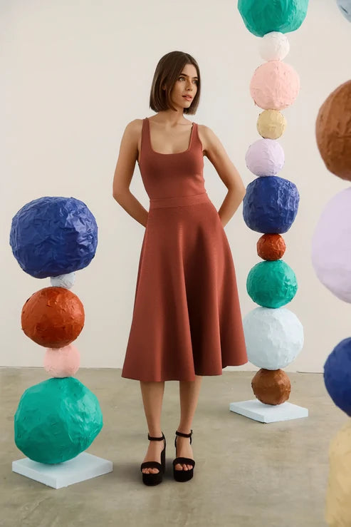 A woman stands in a studio wearing a sleeveless rust-brown set with a square neckline and flared skirt, paired with black platform sandals. She is framed by colorful sculptural paper-ball columns on both sides.