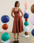 A woman stands in a studio wearing a sleeveless rust-brown set with a square neckline and flared skirt, paired with black platform sandals. She is framed by colorful sculptural paper-ball columns on both sides.