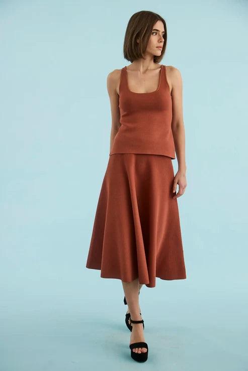 A woman modeling a rust-colored two-piece knit set consisting of a sleeveless scoop-neck top and a matching flared midi skirt, paired with black platform sandals, against a solid light blue background.