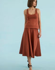 A woman modeling a rust-colored two-piece knit set consisting of a sleeveless scoop-neck top and a matching flared midi skirt, paired with black platform sandals, against a solid light blue background.