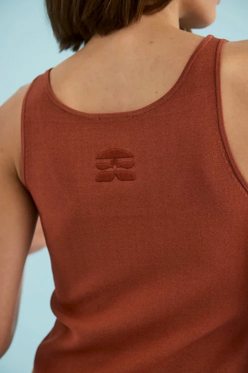 Close-up back view of a person wearing a rust-colored ribbed tank top with a tonal embroidered logo on the upper back, against a light blue background.