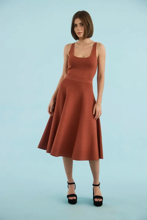 A woman modeling a sleeveless rust-brown midi dress with a scoop neckline and flared skirt, paired with black platform sandals, against a solid light blue studio background.