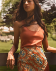 A woman wearing a sleeveless terracotta orange top and a high-waisted orange skirt with blue and beige floral patterns, posing outdoors in a sunny park setting.