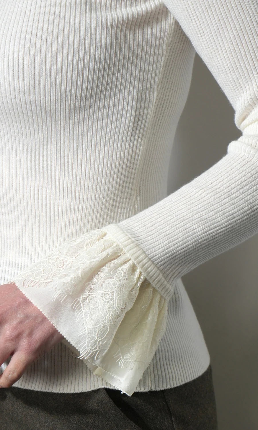 Close-up of a ribbed cream knit top with a flared lace cuff, worn by a model. The image highlights the texture of the cream ribbed fabric and the delicate lace detail on the sleeve cuff.