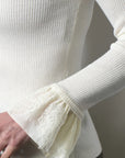 Close-up of a ribbed cream knit top with a flared lace cuff, worn by a model. The image highlights the texture of the cream ribbed fabric and the delicate lace detail on the sleeve cuff.