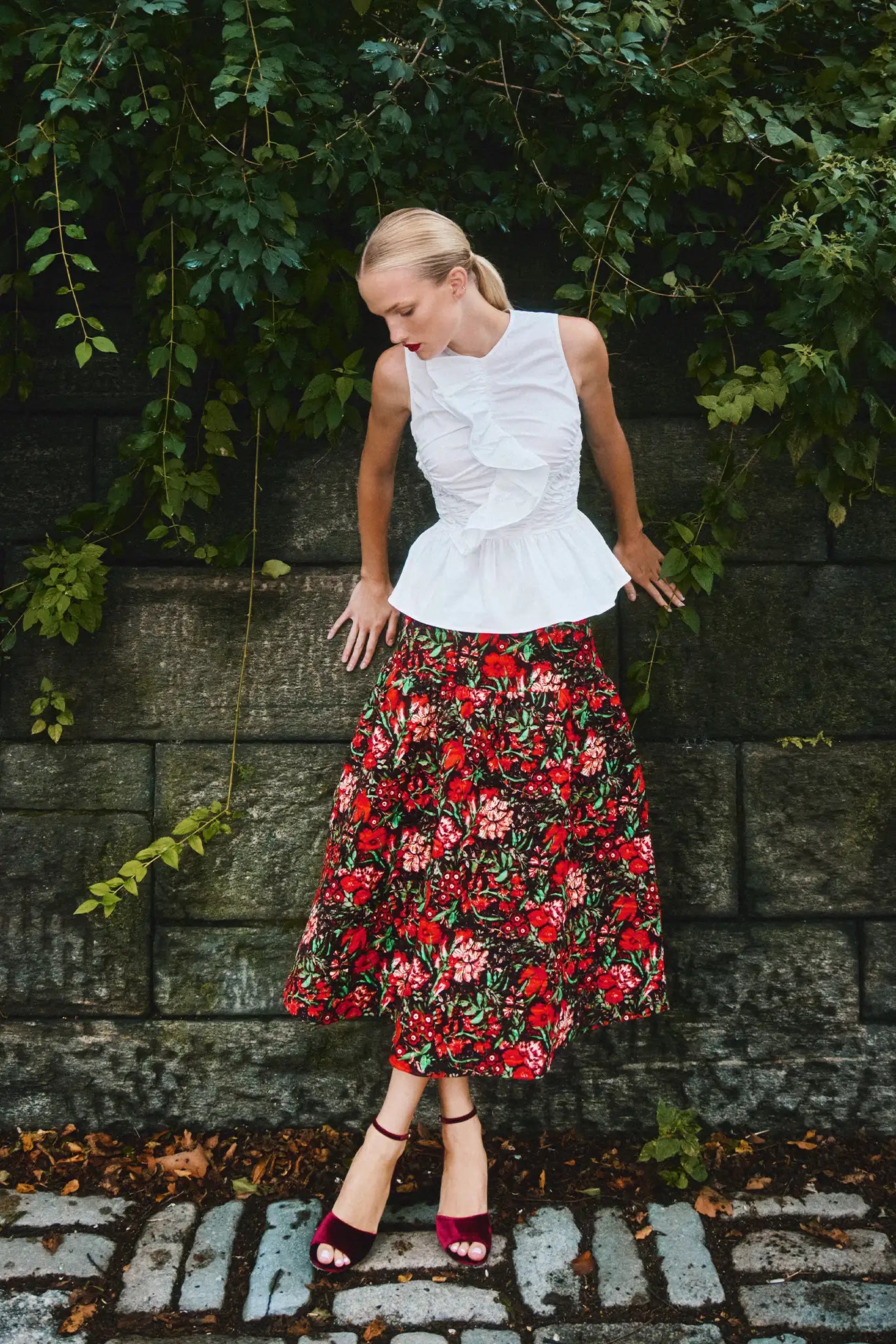 Woman wearing a white top and red floral skirt standing against a stone wall with greenery.