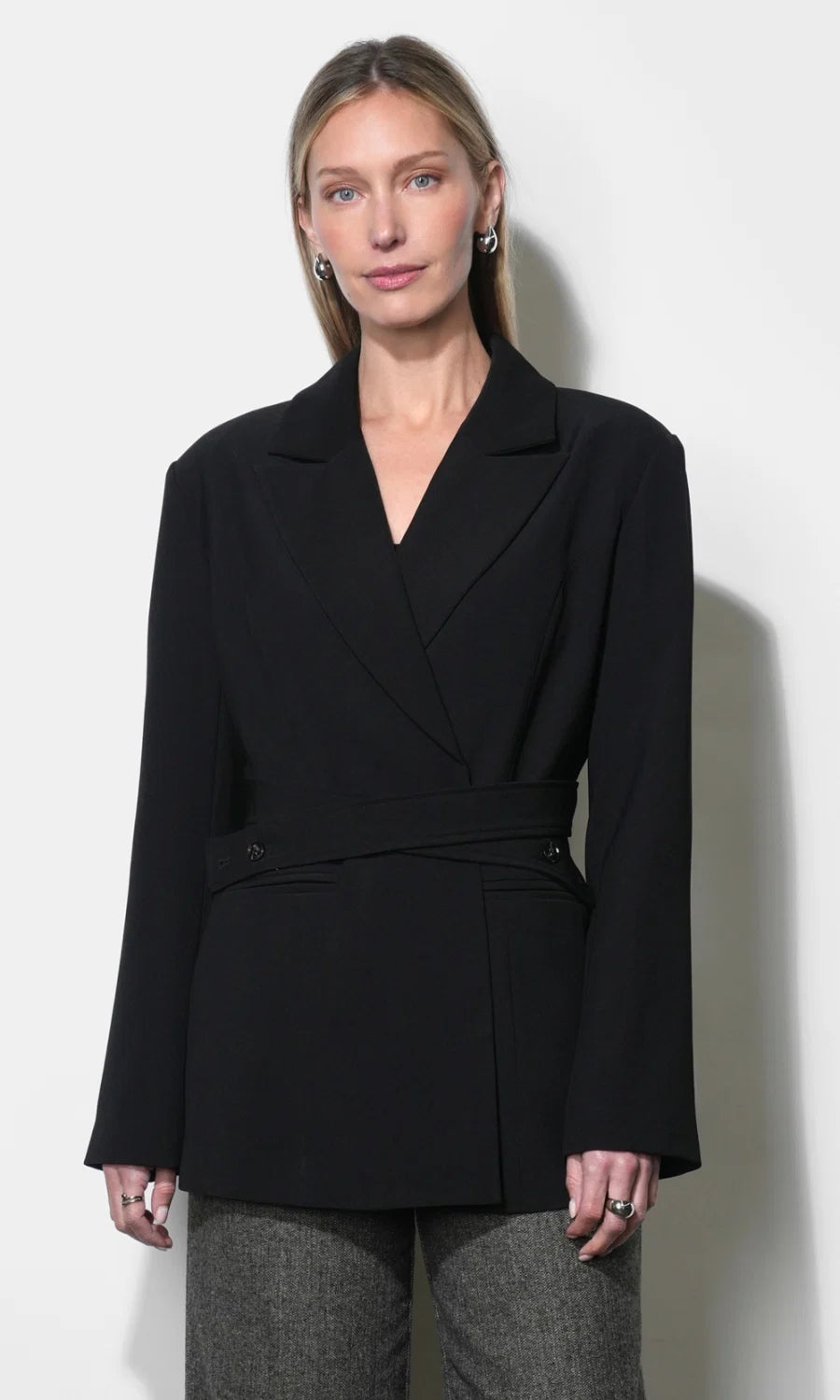 A woman modeling a black wrap-style blazer with a self-tie waist belt, paired with gray textured trousers, against a plain off-white background.