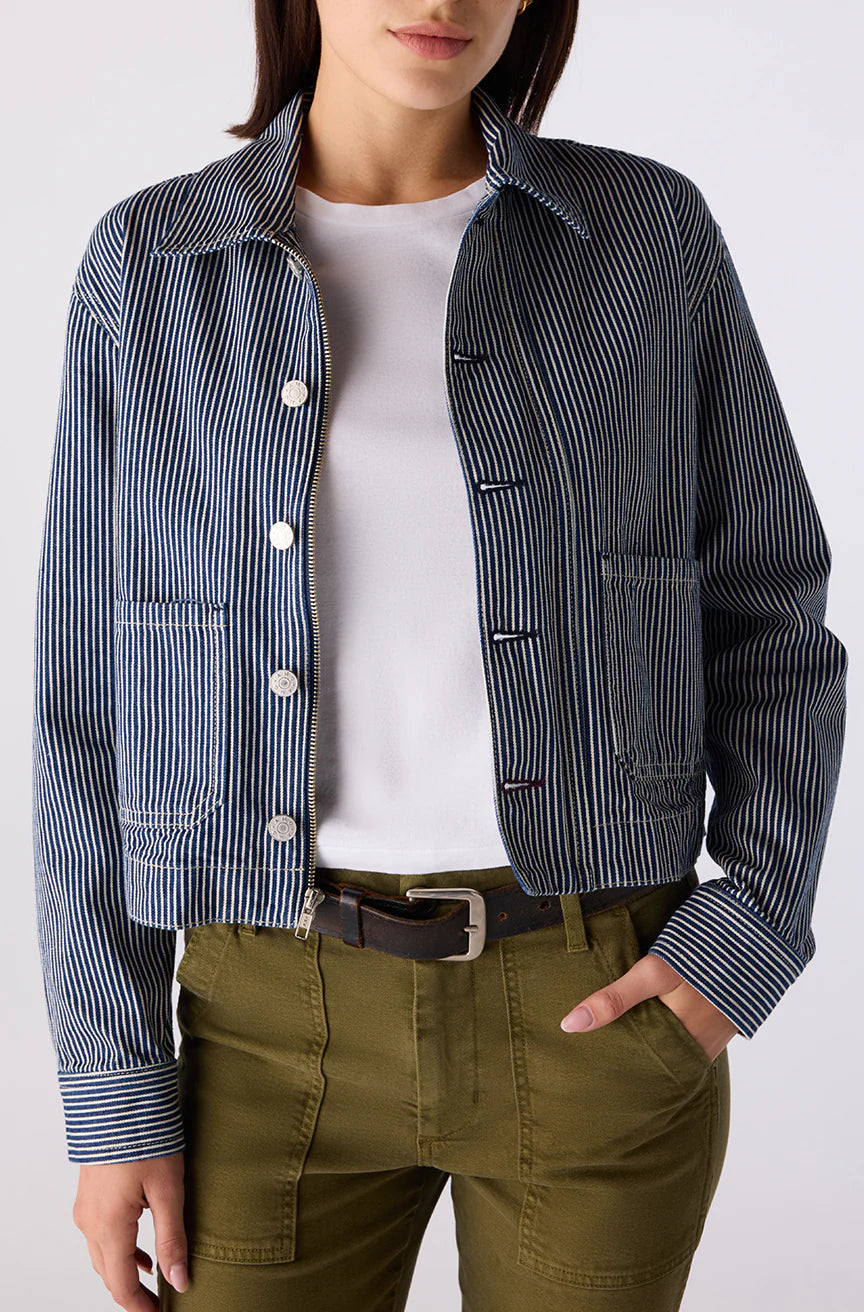 Person wearing a striped jacket over a white shirt with green pants on a plain background